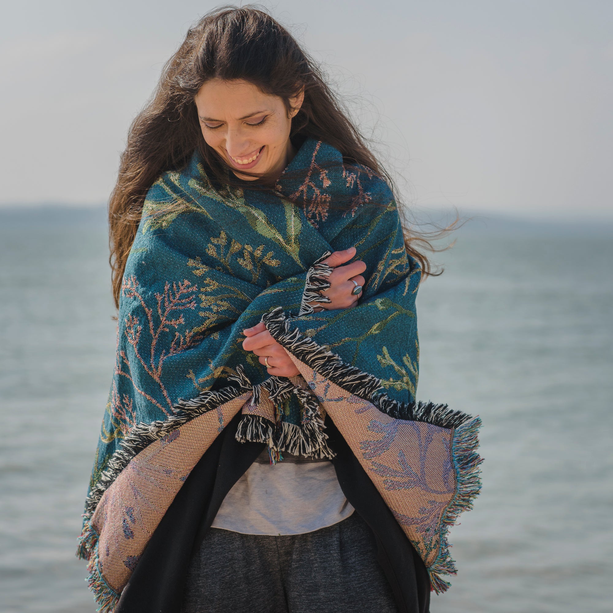 delicate seaweed design on woven wrap blanket with fringe wrapped around womans shoulders with the sea in the background