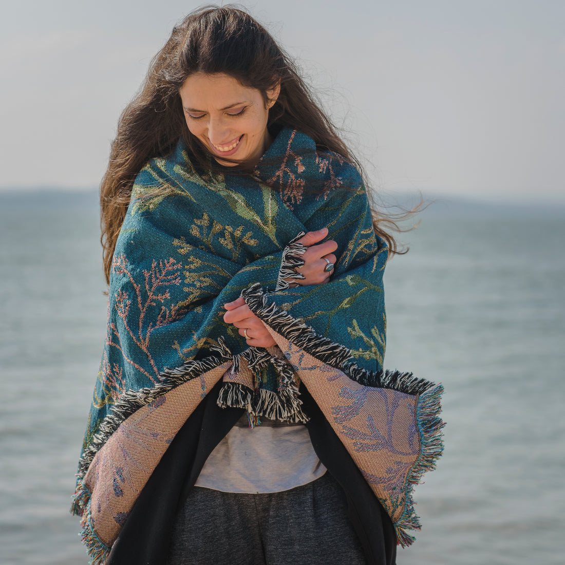 delicate seaweed design on woven wrap blanket with fringe wrapped around womans shoulders with the sea in the background