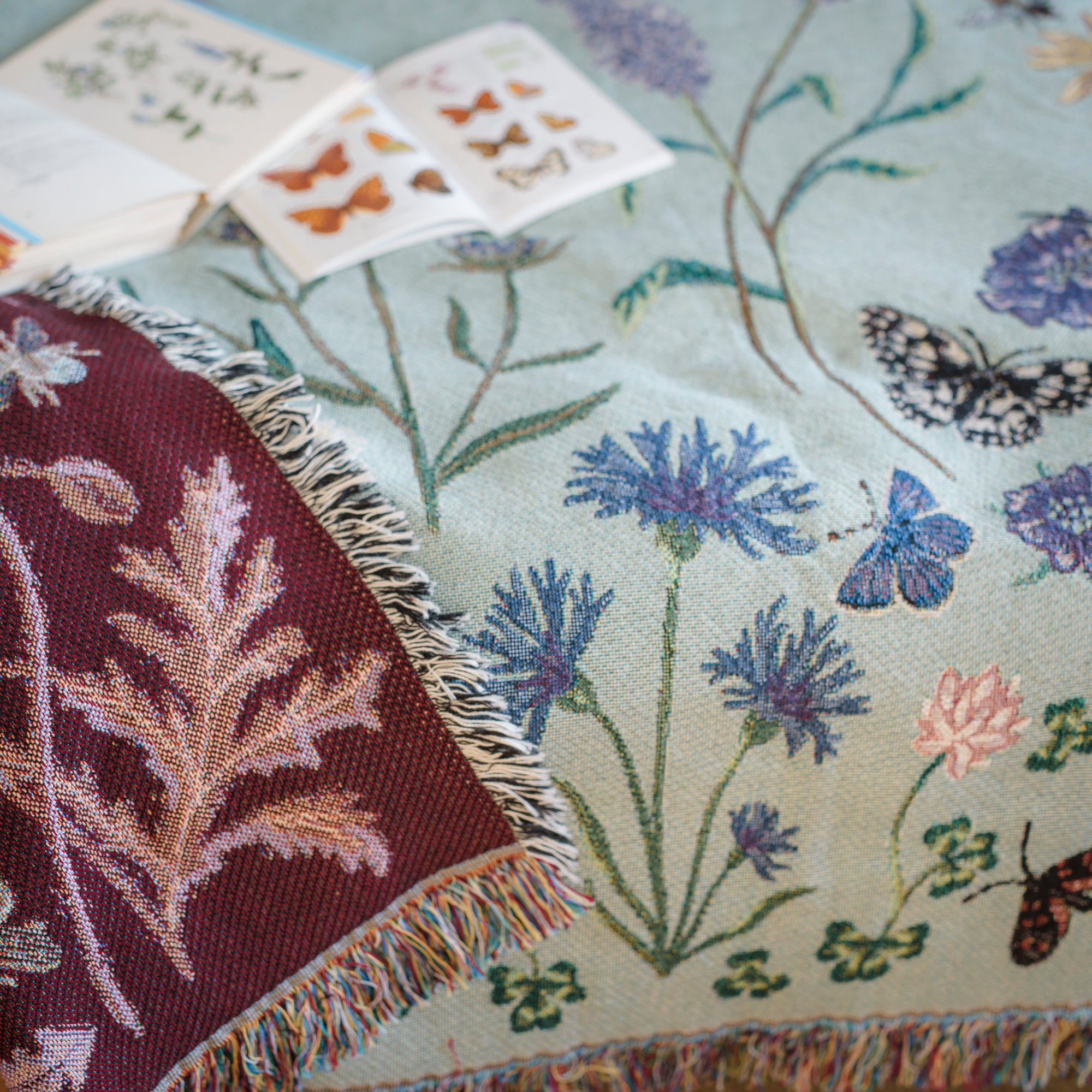 pale sage green woven blanket with ruby red reverse with details of butterflies and wild flowers, showing fringe and woven details