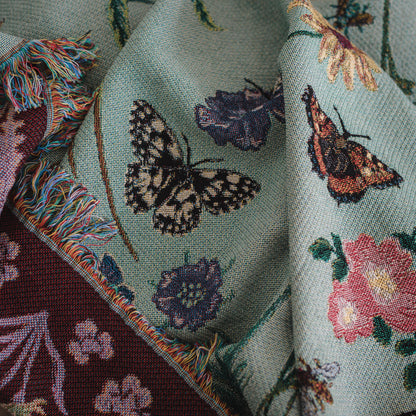 pale sage green woven blanket with ruby red reverse with details of butterflies and wild flowers, showing fringe and woven details