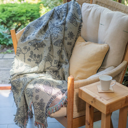 Pretty grey blanket with black pattern of moths, berries, leaves and flowers, draped over a chair in a convervatory with sunlight