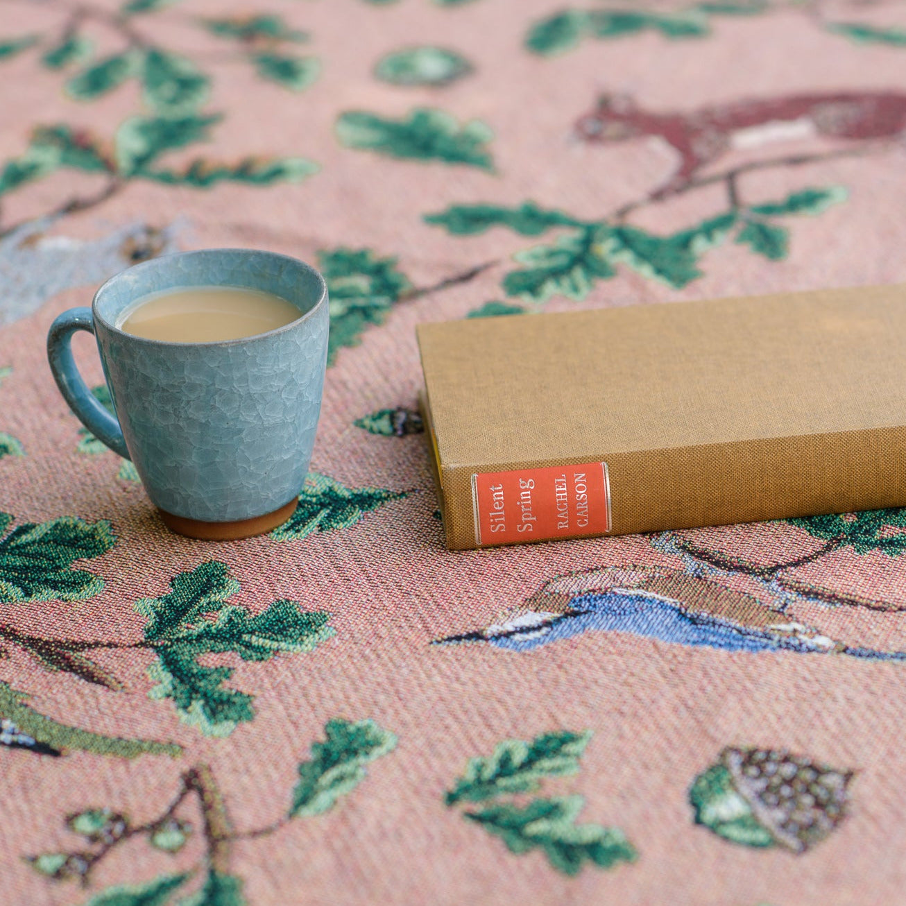 russet coloured woven blanket with teal reverse featuring birds, squirrels and oak, with tea cup and book