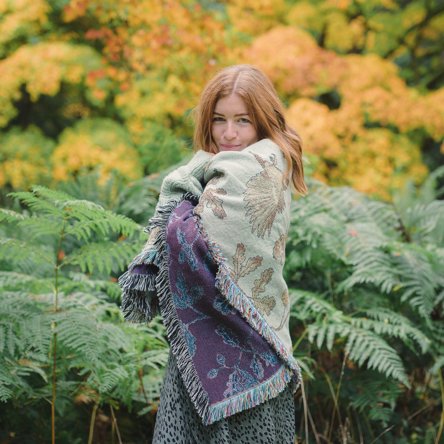 A woman standing in a forest with an Arcana Wild Oak Wrap Blanket wrapped around her.